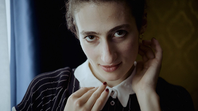 niki blau image 05 Close-up of the Escort Niki Blau. Her face dominates the frame, her hands framing it: One resting on her chin, the other at her ear. With widened, glistening eyes, she gazes seductively upward into the camera. Her expression appears both innocent and calculating. Her mouth is slightly open, her lips glistening. Her dress resembles that of a maid.