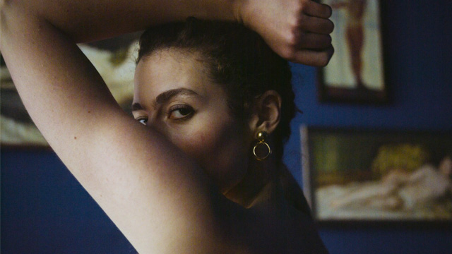 niki blau image 08 Seitlicher Close-up von Nikis Gesicht, das halb von über ihren Kopf gebeugten Armen verdeckt ist. Ihr Blick ist über die Schulter in die Kamera gerichtet. Sie ist dezent geschminkt und trägt auffällige goldene Ohrringe.
