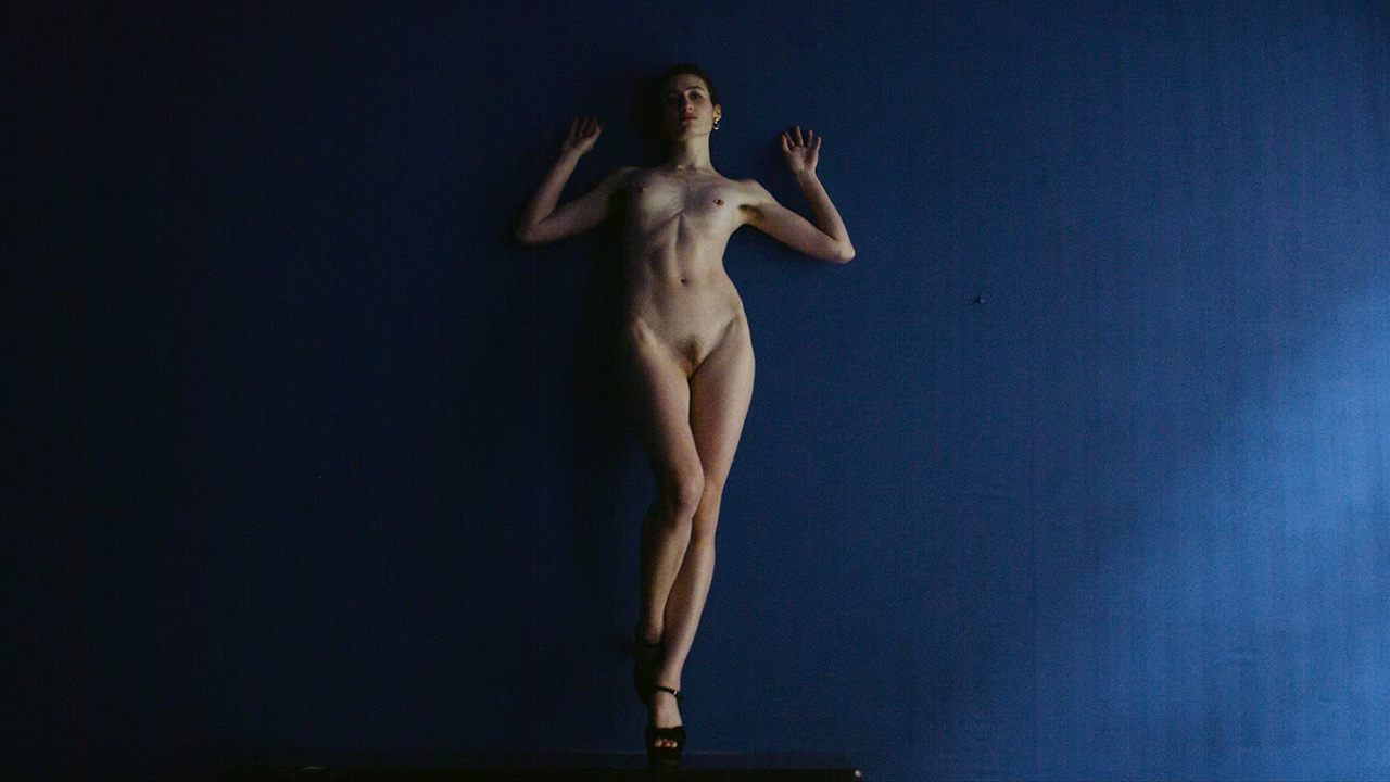 niki blau image 10 Niki steht vollkommen nackt vor einer blauen Wand. Ein Bein gestreckt, das andere angewinkelt, balanciert sie auf einem Kaminsims, den wir nur im Anschnitt erahnen können, den Körper leicht an die Wand gelehnt. Ihre Arme und Hände ruhen angewinkelt an der Wand. Durch Haltung und Lichteinfall erscheint ihr geschmeidiger Körper zugleich schlank und kurvig, ihre kleinen Brüste rund und voll. Ihr Blick trifft die Kamera unverhohlen und direkt.