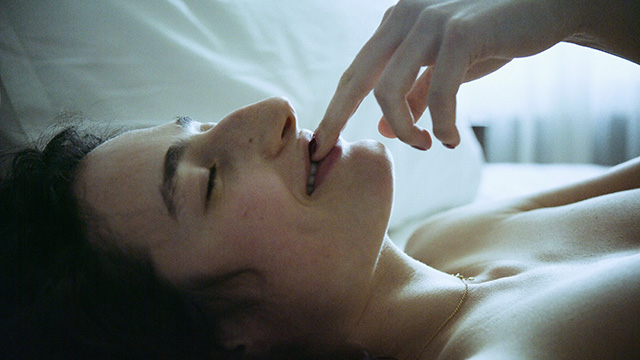 niki blau image 18 Zu sehen ist ein Close-up von Nikis Profil. Sie lächelt und hält dabei zwei Finger in ihren leicht geöffneten Mund. Das Licht durch das Fenster betont ihre zarte Haut.