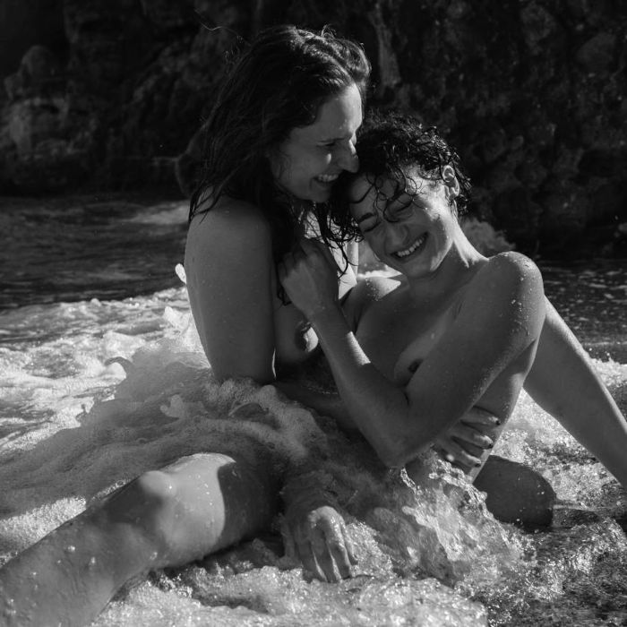 luisa niki Luisa und Niki sitzen lachend nackt sich in den Armen haltend am Meeresufer, Gischt einer frischen Welle umschließt sie.