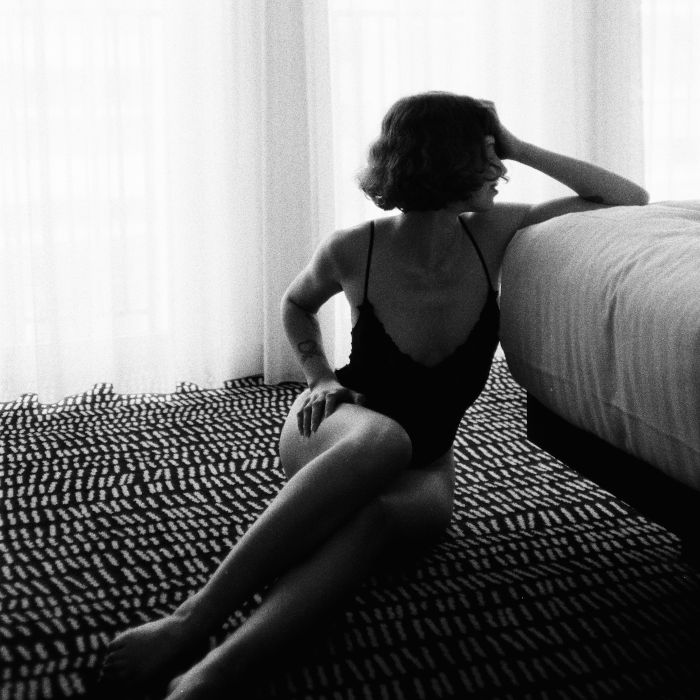 matilda niki In einen Body gekleidet, sitzt Matilda auf dem gemusterten Teppichboden eines Hotelzimmers. Die Beine leicht angewinkelt, ruht ein Arm auf ihren Knien, der andere auf dem Bett – mit der Hand stützt sie sanft die Stirn. Ihr Gesicht ist von der Kamera abgewandt.