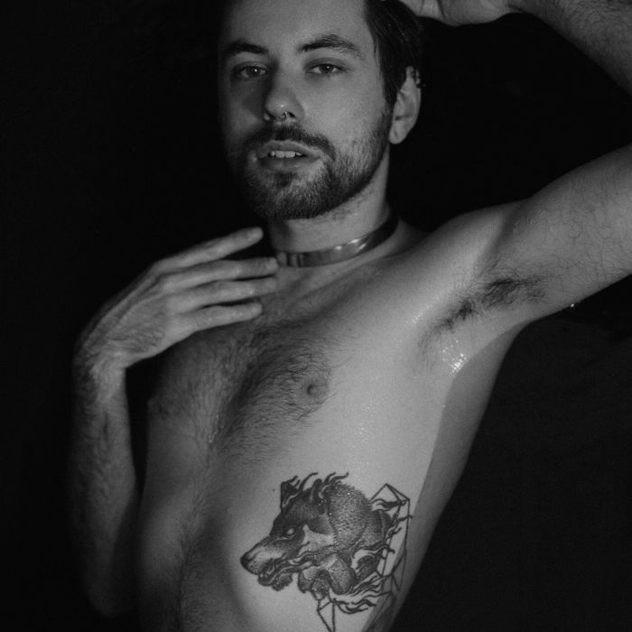 dante niki Wir schauen von oben auf Dante, der im Wasser zu liegen scheint. Eine Hand streichelt seinen Hals, die andere ruht hinter seinem Kopf, sein Blick in die Kamera gerichtet. Auf seinem leicht behaarten Oberkörper ist ein Tattoo eines Wolfes zu erkennen.